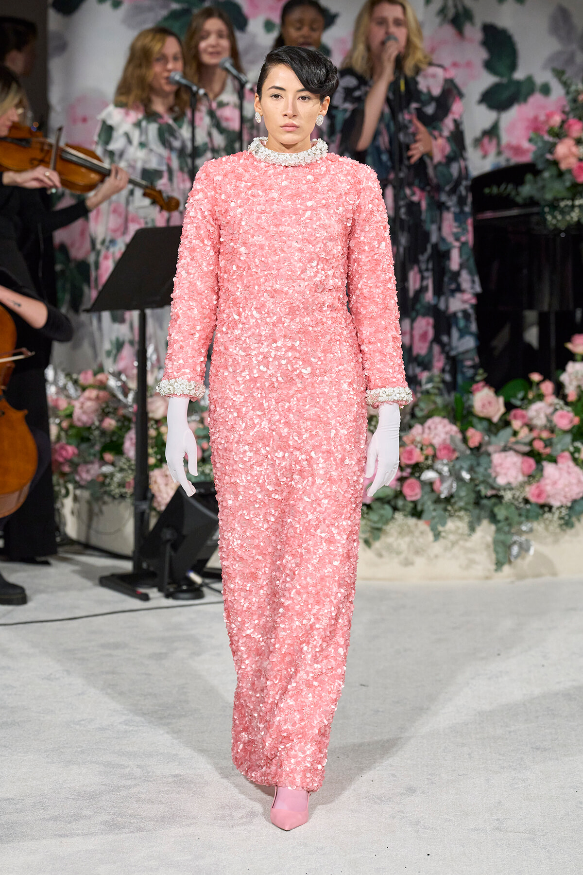 Woman model walking the runway in a pink sequin gown with white gloves and pink shoes, floral backdrop behind her.