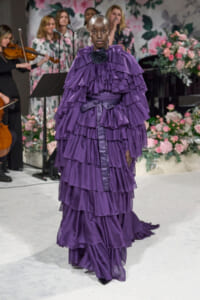 Model walking a runway in a voluminous, layered purple ruffled gown against a floral backdrop.