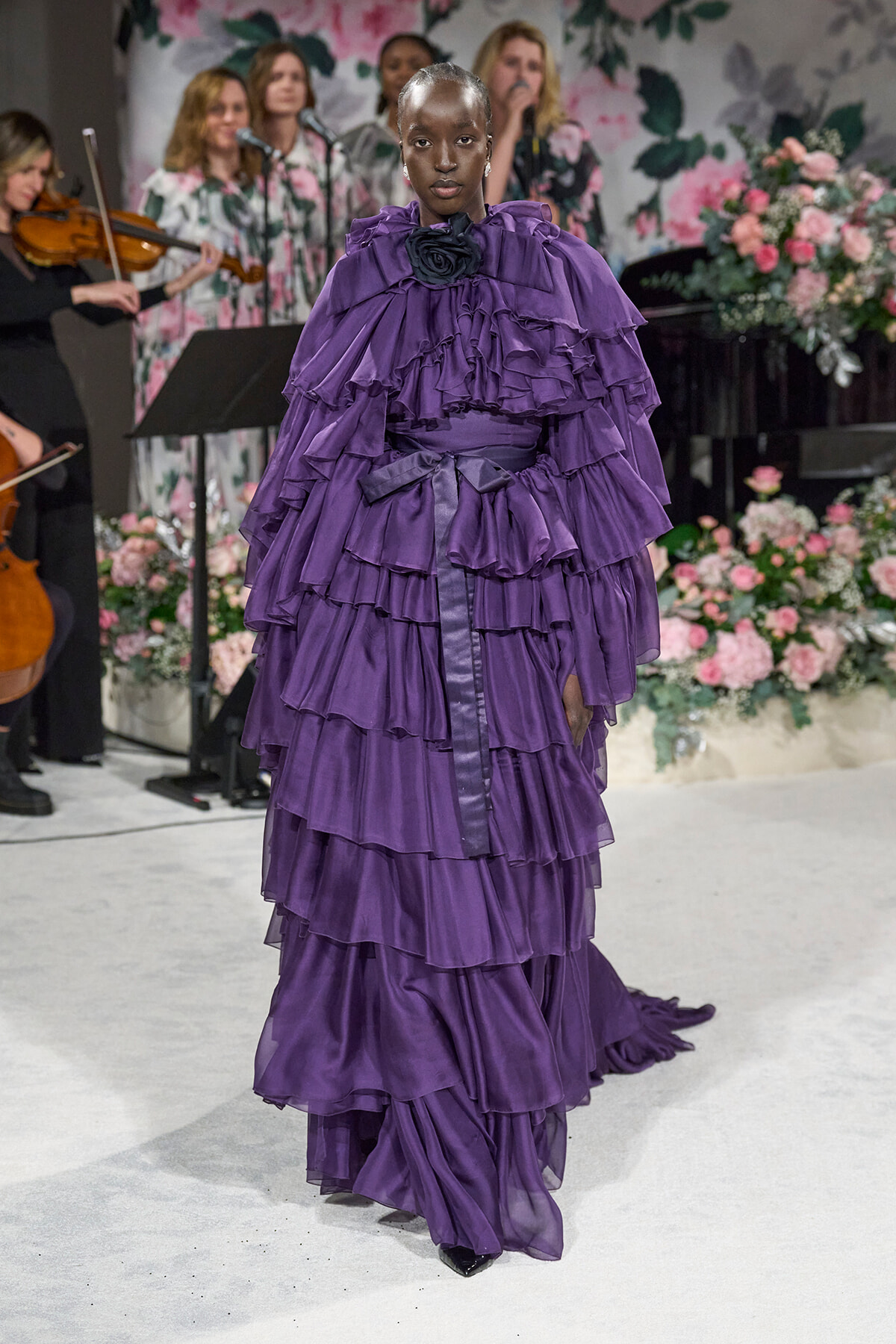Model walking a runway in a voluminous, layered purple ruffled gown against a floral backdrop.