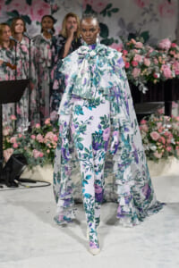Model walks a floral-print gown with layered ruffles and a bow at the neck on a flower-filled runway backdrop.