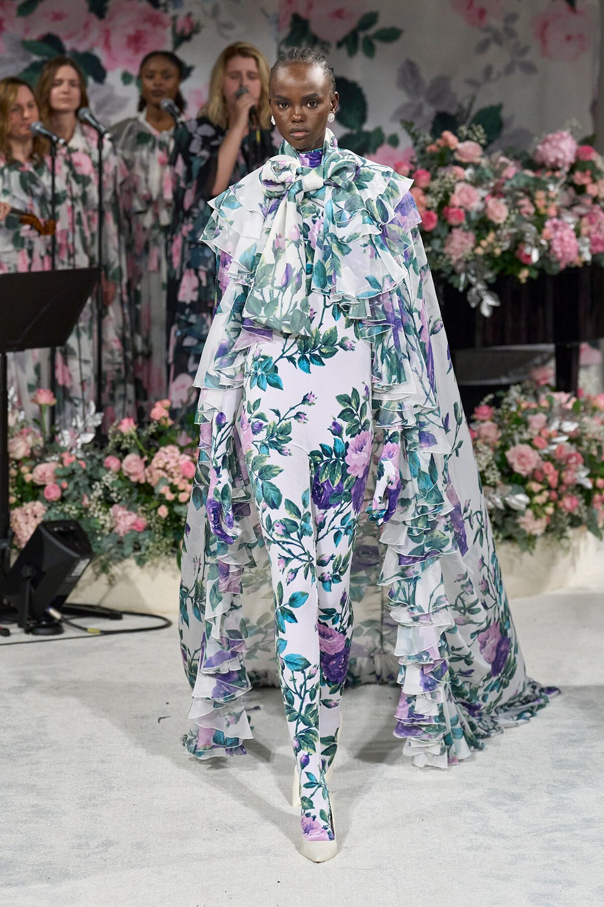 Model walks a floral-print gown with layered ruffles and a bow at the neck on a flower-filled runway backdrop.