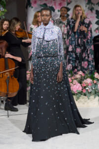 Model walks in a long black floral gown with a large bow at the neck, white and purple blossoms, on a runway with a floral backdrop and musicians in the background