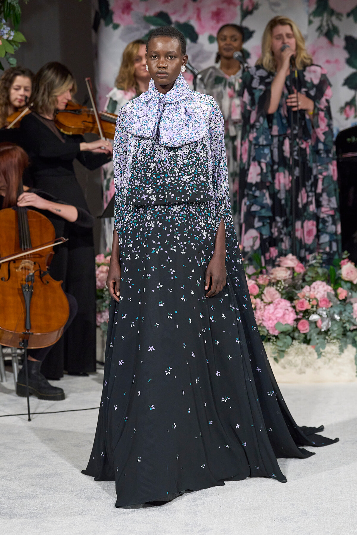 Model walks in a long black floral gown with a large bow at the neck, white and purple blossoms, on a runway with a floral backdrop and musicians in the background