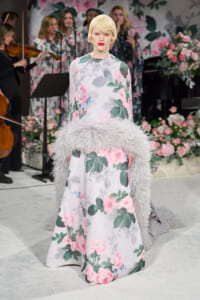 Model on a runway wearing a pastel floral gown with a fluffy gray feather stole, flowers backdrop, and musicians in the background