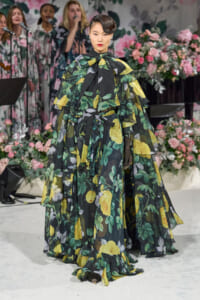 Model wearing a voluminous sheer floral gown with green leaves and yellow flowers on a runway backdrop of pink flowers and singers in background.