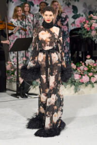 Model wearing a long sheer floral gown with a black bow, feather-trimmed cuffs, and bold lipstick on a floral-runway backdrop with singers in the background.