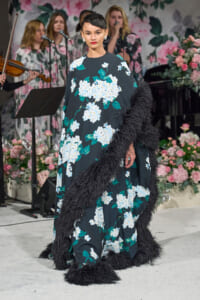 Model on a runway wearing a long teal floral gown with white blossoms and a black furry trim, in front of a floral stage backdrop.