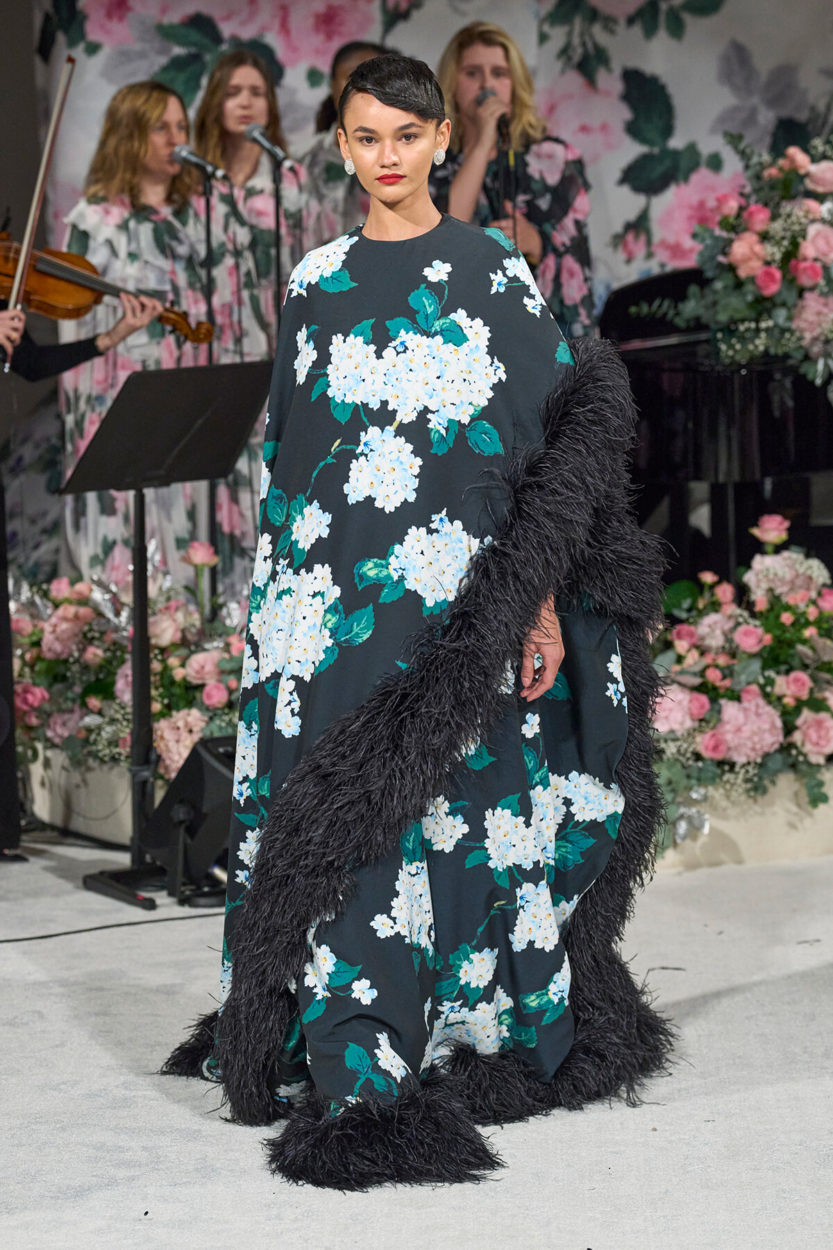 Model on a runway wearing a long teal floral gown with white blossoms and a black furry trim, in front of a floral stage backdrop.