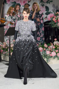 Model in a black floral-embellished gown walks the runway with long black gloves and a voluminous train, backdrop of pink flowers and a singing group behind.