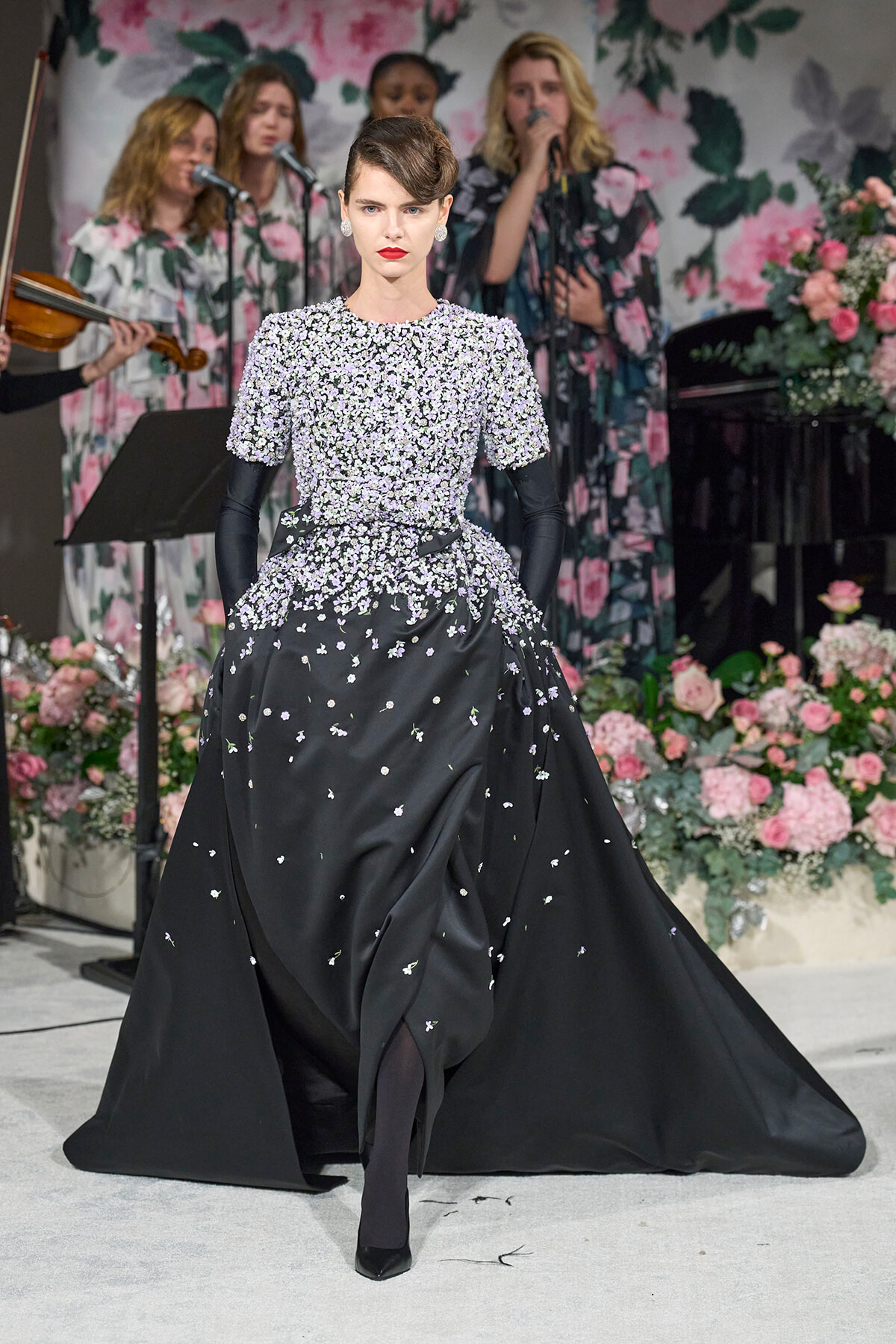Model in a black floral-embellished gown walks the runway with long black gloves and a voluminous train, backdrop of pink flowers and a singing group behind.