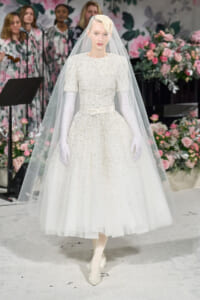 Runway model in a pearl-embellished white wedding gown with a long veil and white gloves, surrounded by pink flowers in a fashion show backdrop.