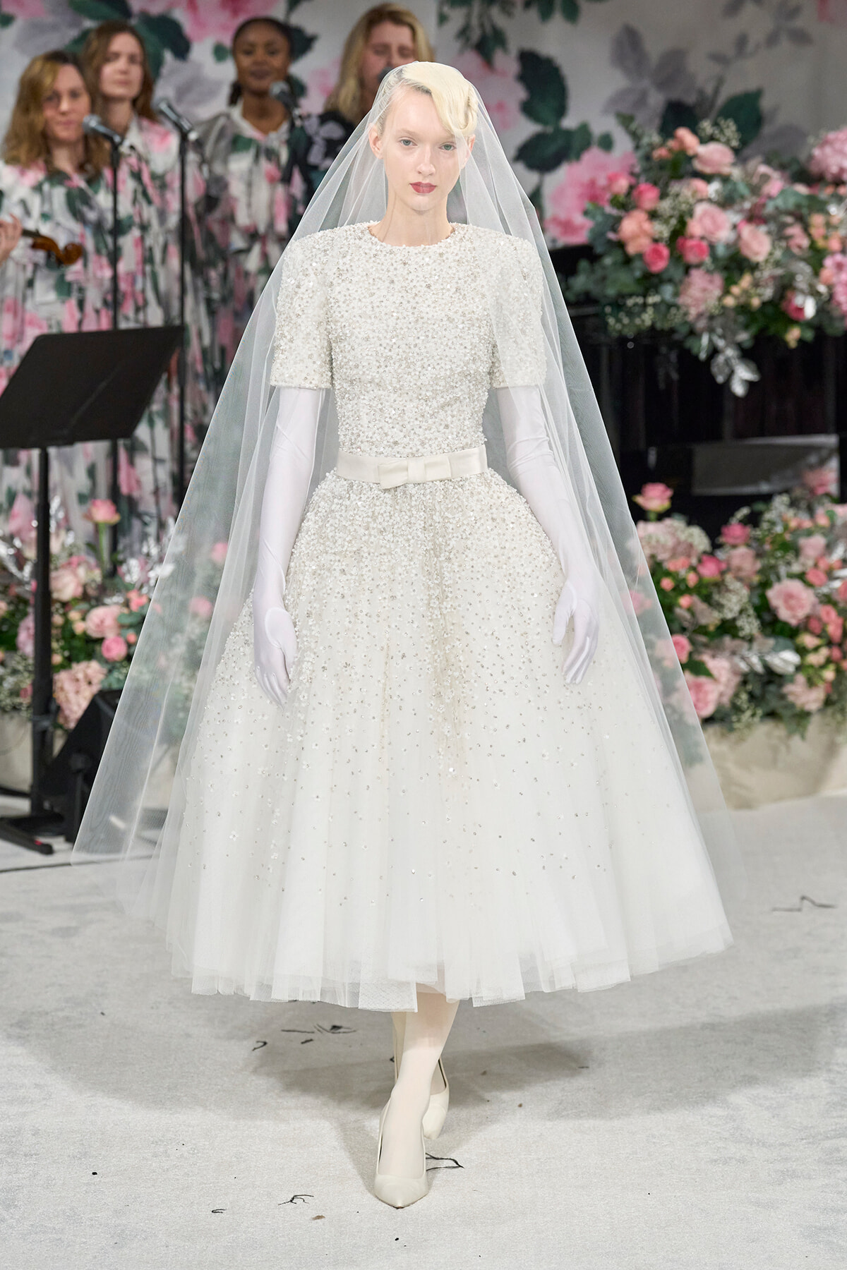 Runway model in a pearl-embellished white wedding gown with a long veil and white gloves, surrounded by pink flowers in a fashion show backdrop.