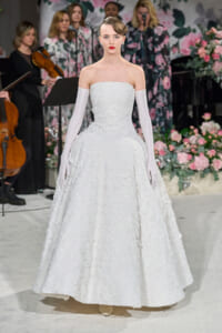 Model wearing a strapless white lace wedding gown with long white gloves, standing on a floral runway with musicians in the background.