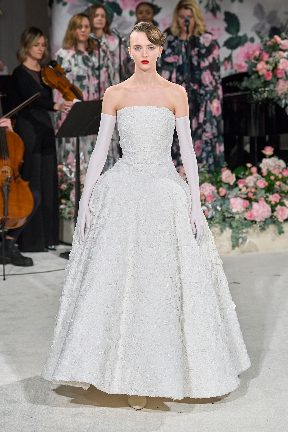 Model wearing a strapless white lace wedding gown with long white gloves, standing on a floral runway with musicians in the background.
