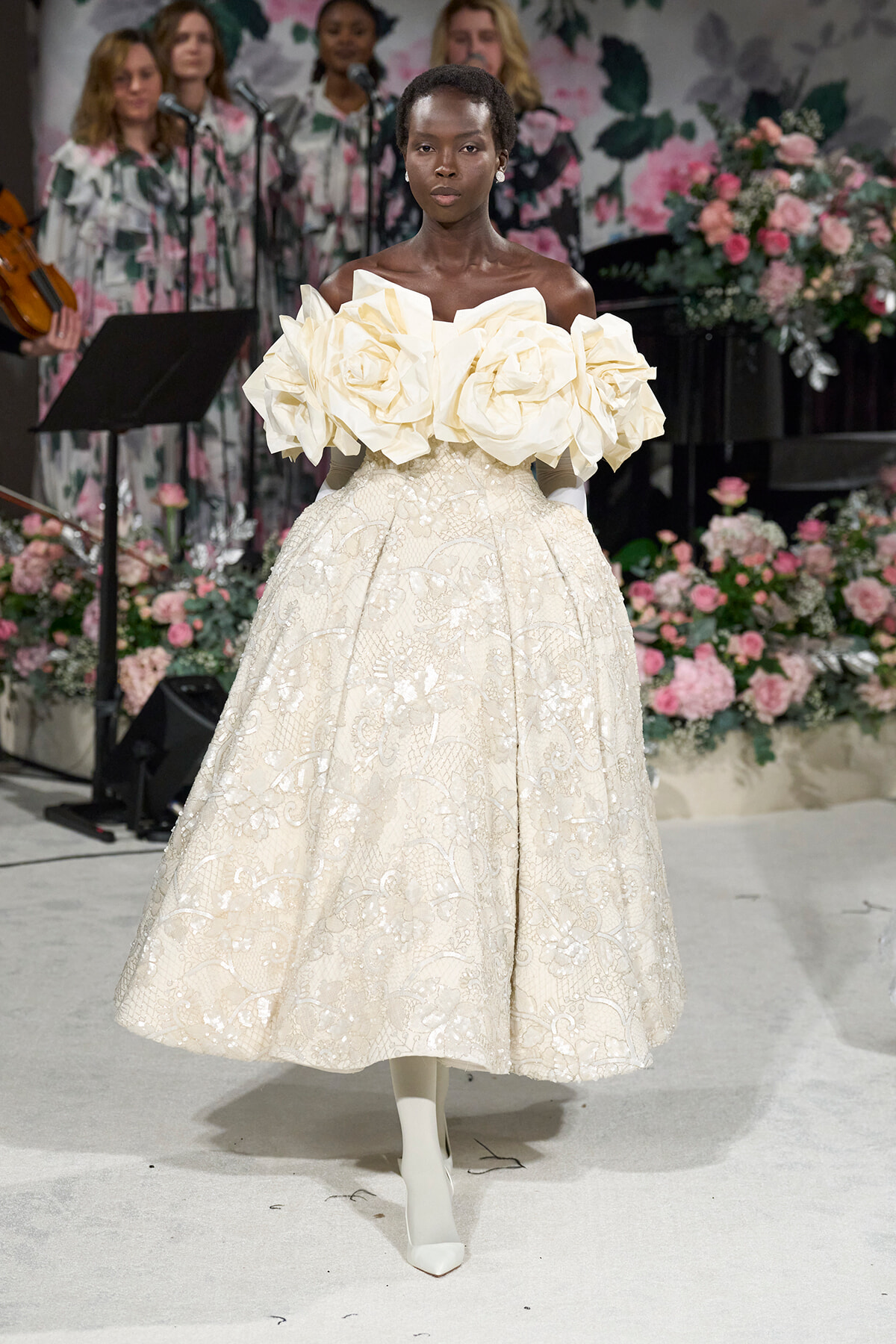 Model on a runway wearing an ivory ball gown with oversized floral rosette shoulders and a lace skirt, against a floral backdrop.
