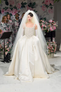 Bride model in an ivory ball gown with a jeweled bodice and flowing veil on a floral runway backdrop of pink blooms.