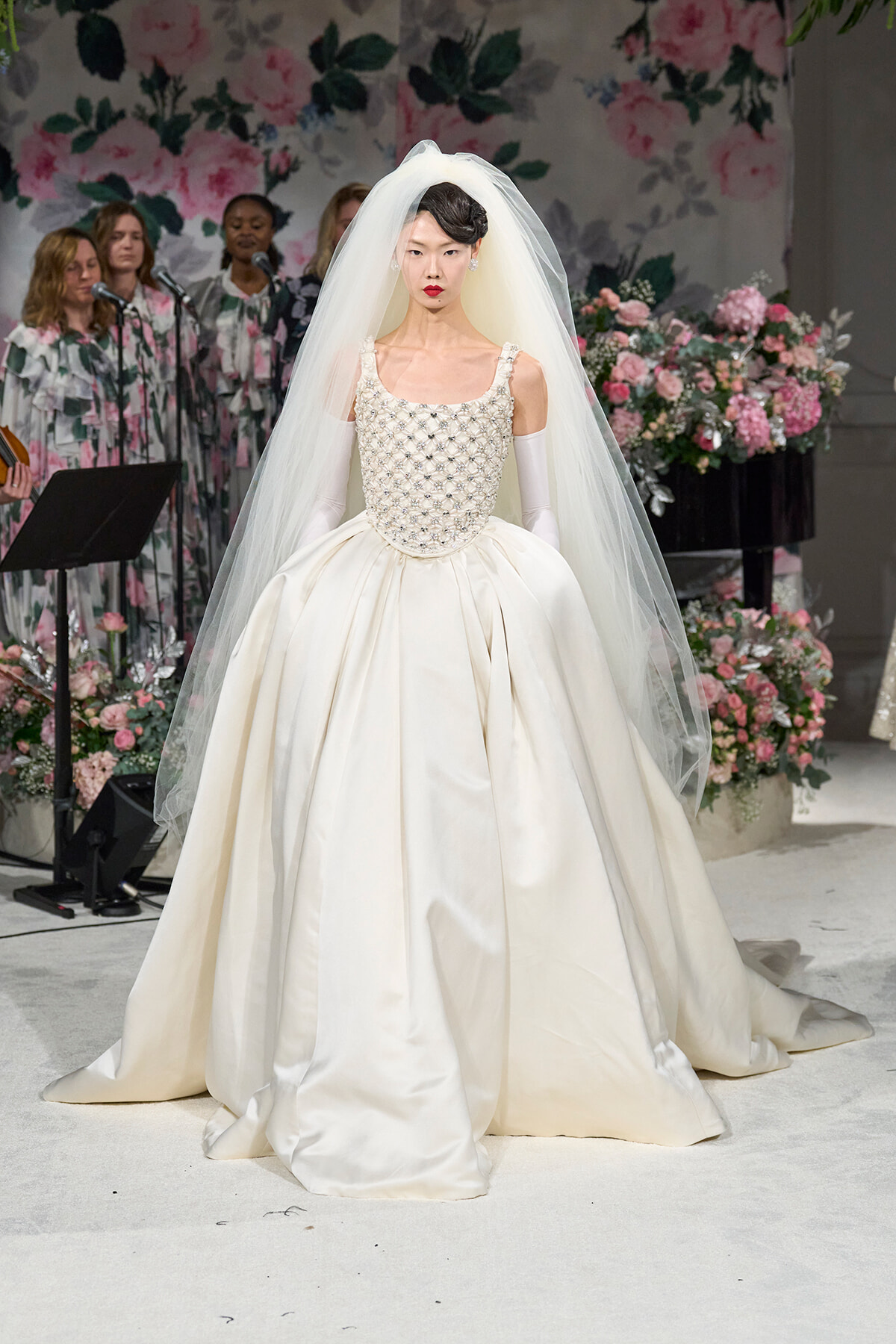 Bride model in an ivory ball gown with a jeweled bodice and flowing veil on a floral runway backdrop of pink blooms.