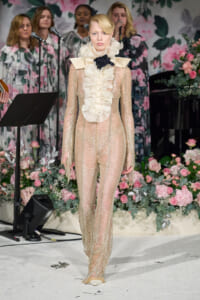Model walks the runway in a cream ruffled high-neck gown with sheer beaded sleeves against a floral backdrop.