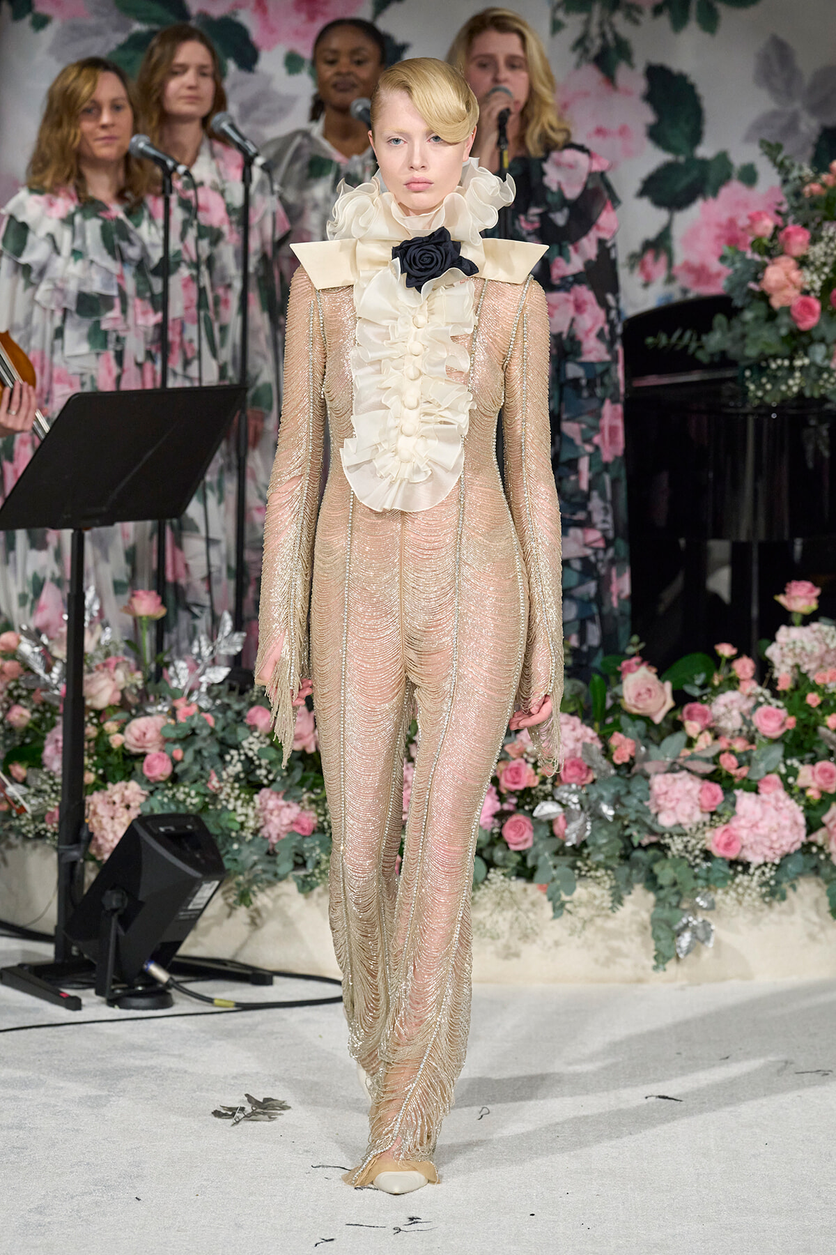 Model walks the runway in a cream ruffled high-neck gown with sheer beaded sleeves against a floral backdrop.