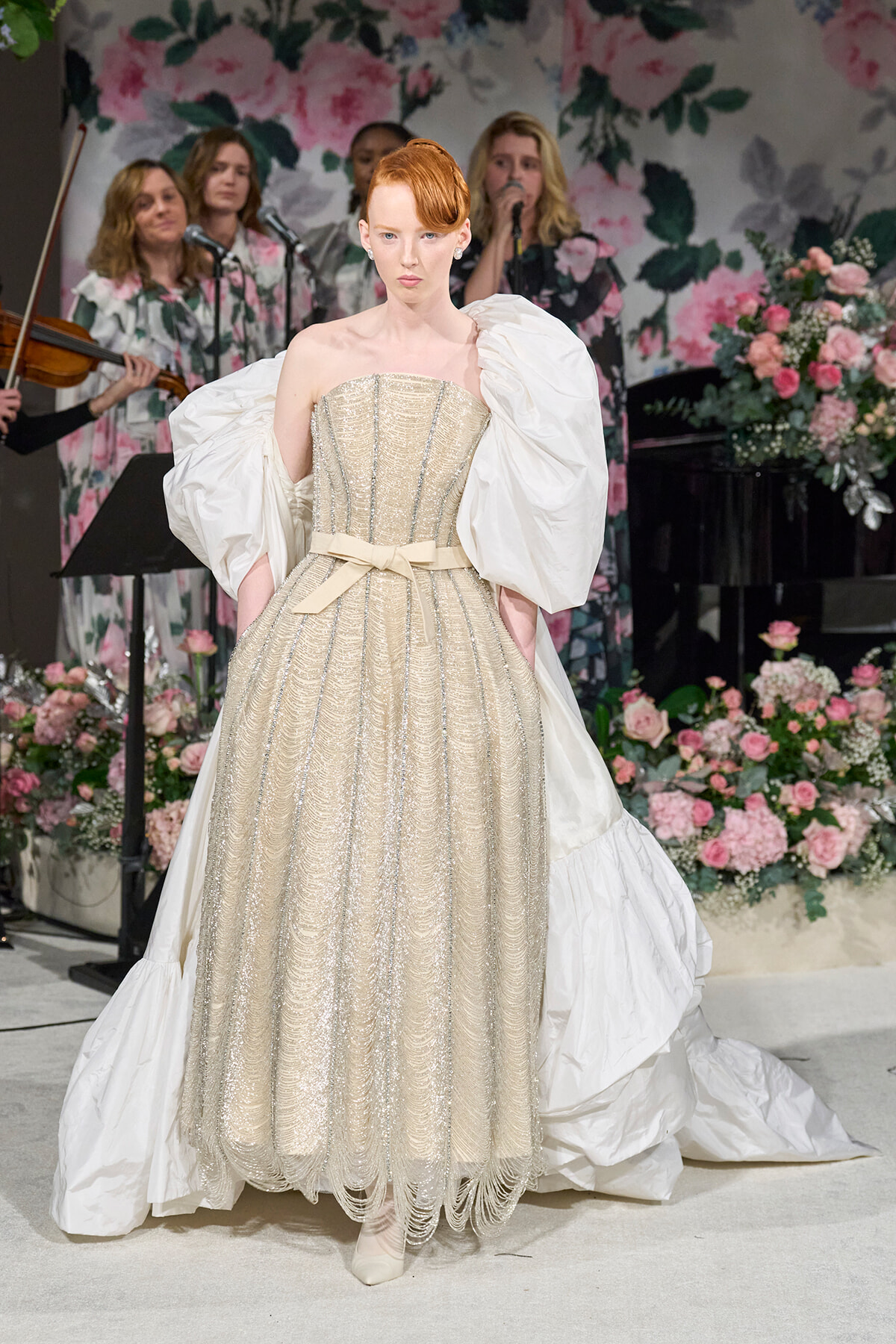 Model wears a gold, beaded ballgown with a large white cape and pale ribbon belt on a floral-stage runway.