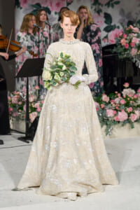 Bride in an ivory lace wedding gown holds a white bouquet, standing on a floral-stage backdrop with musicians in the background.