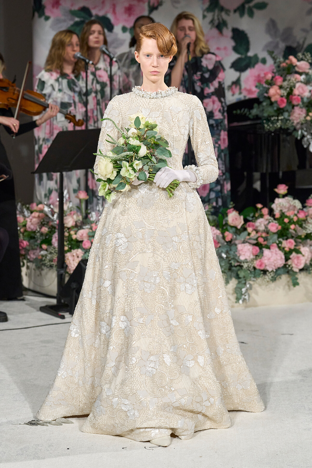 Bride in an ivory lace wedding gown holds a white bouquet, standing on a floral-stage backdrop with musicians in the background.