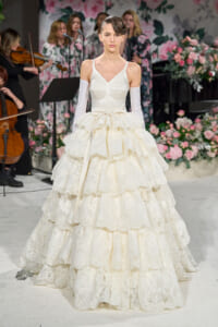 Model walking a floral-themed runway in a voluminous ivory lace ball gown with off-the-shoulder sleeves and a bow at the waist.