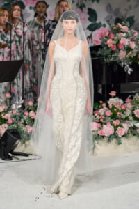 Model wearing an embellished white wedding gown with a long veil on a floral fashion-show runway.
