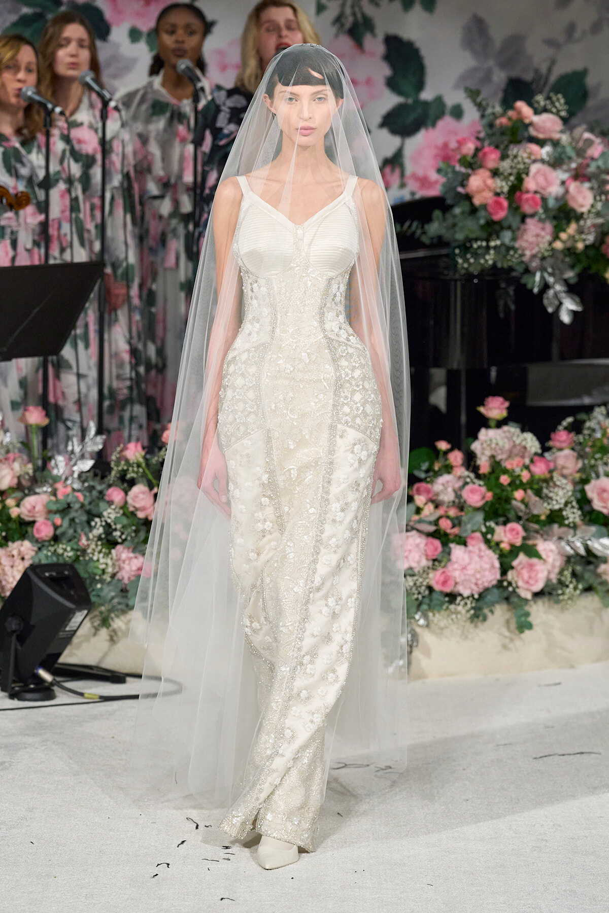 Model wearing an embellished white wedding gown with a long veil on a floral fashion-show runway.
