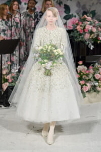 Bride in a sparkling white gown and veil holding a white rose bouquet, standing among pink floral decorations.