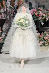 Bride in a pearl-beaded white wedding gown and veil, holding a white rose bouquet, on a floral backdrop.