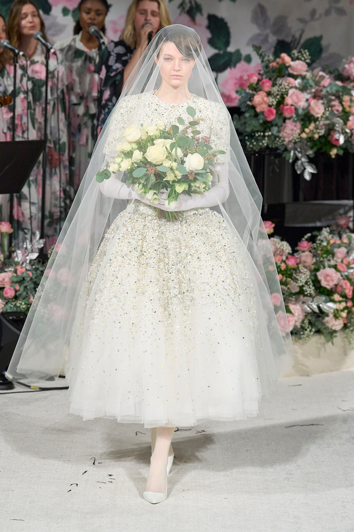 Bride in a pearl-beaded white wedding gown and veil, holding a white rose bouquet, on a floral backdrop.