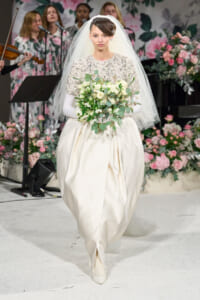 Bride in an ivory embellished gown with a white veil, carrying a white bouquet, walking down a floral runway.