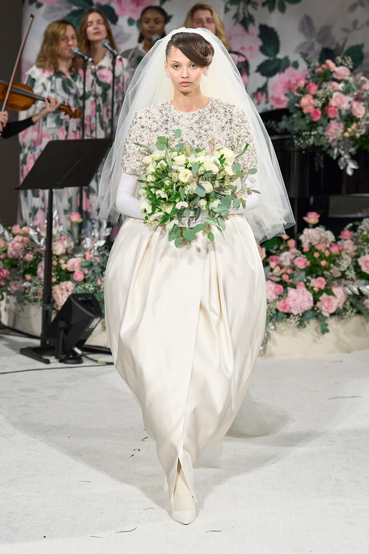 Bride in an ivory embellished gown with a white veil, carrying a white bouquet, walking down a floral runway.