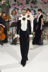 Model walks the runway in a black velvet gown with a white ruffled bib and cuffs, accented by a black rose brooch, against a floral backdrop.