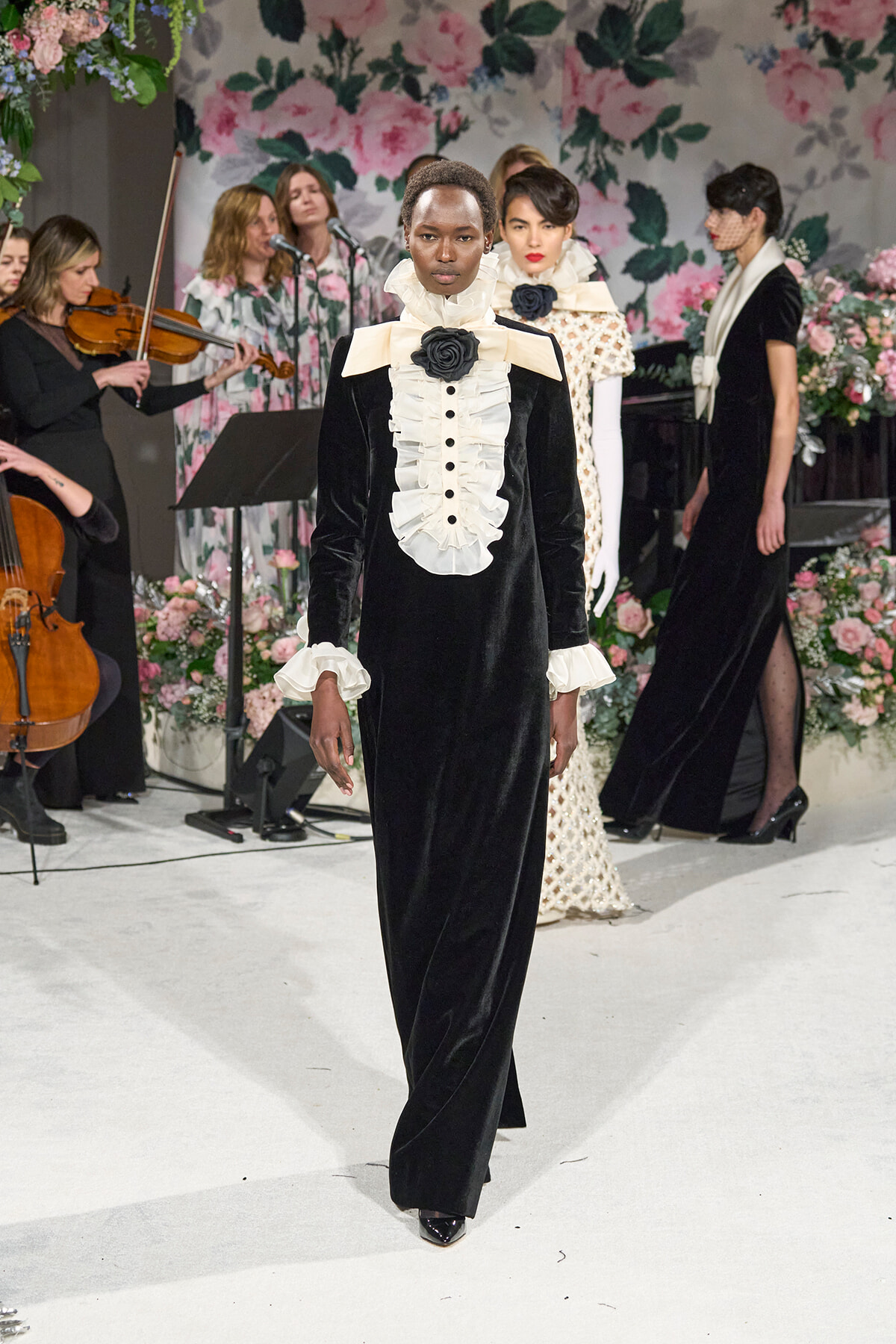 Model walks the runway in a black velvet gown with a white ruffled bib and cuffs, accented by a black rose brooch, against a floral backdrop.
