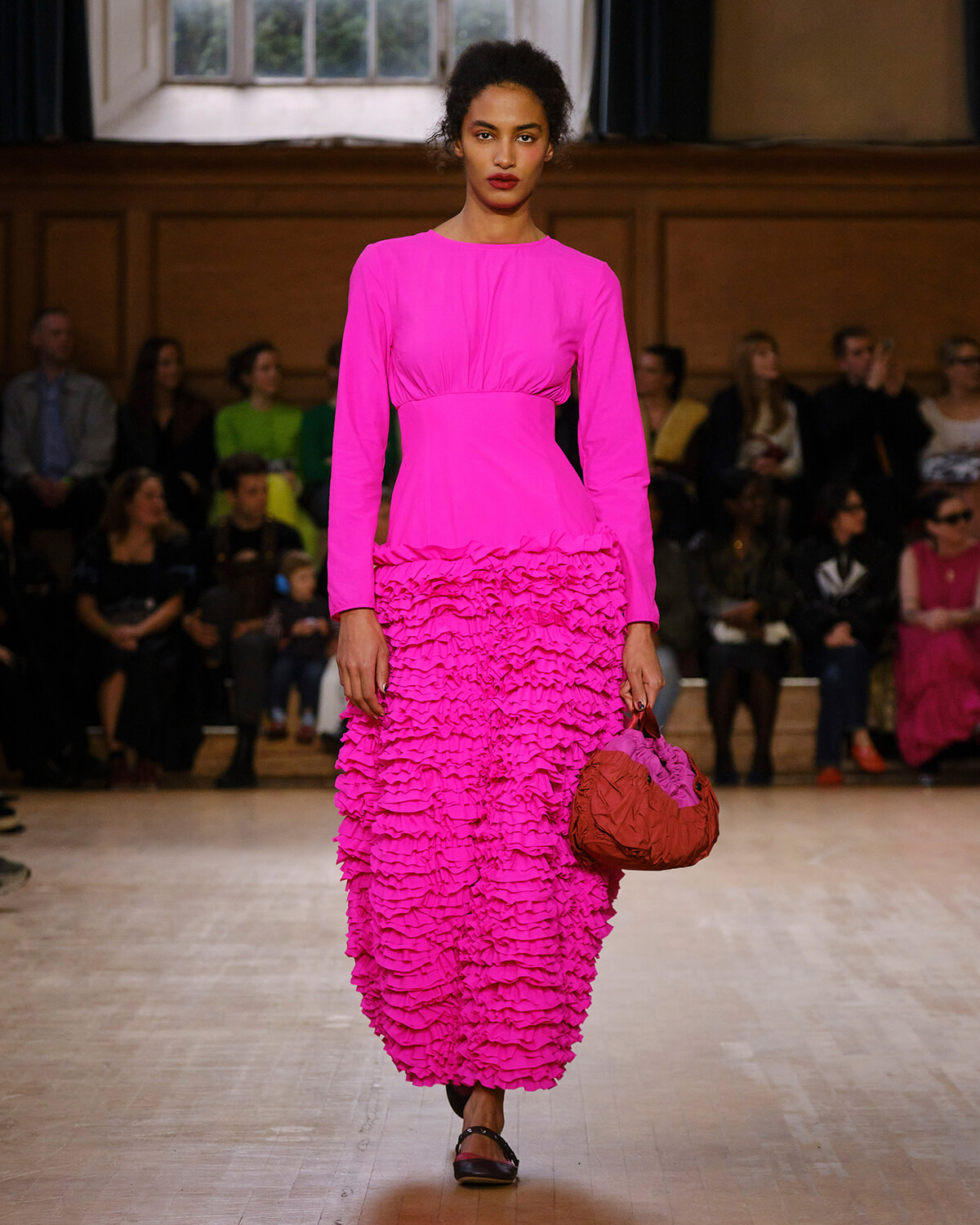 Model on runway wearing a bright pink long-sleeve top and tiered ruffled pink midi skirt, carrying a pink and brown handbag; audience watches in the background.
