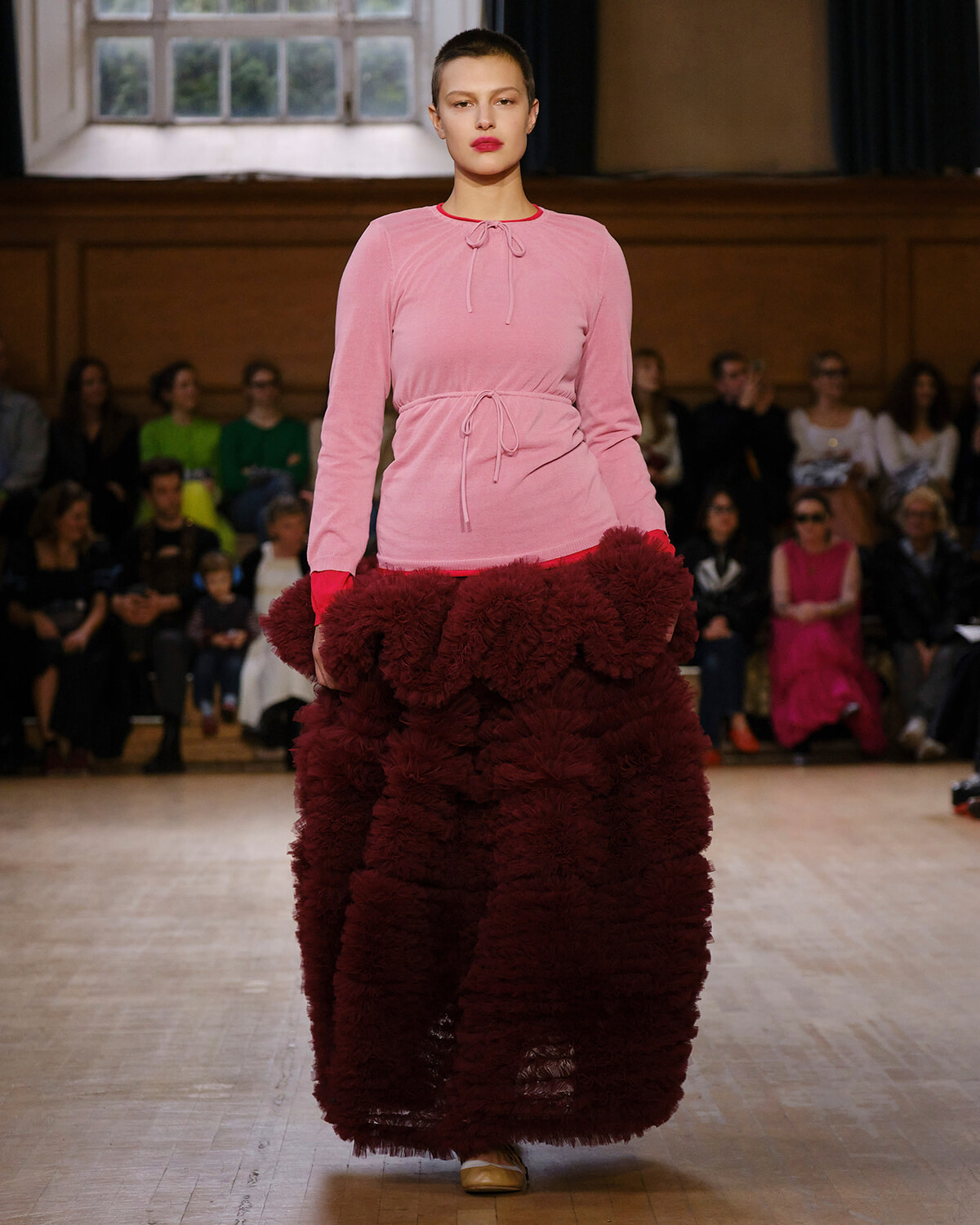 Runway model in a pale pink knit top with a tied waist and a dramatic burgundy fluffy skirt; audience watches from the background.