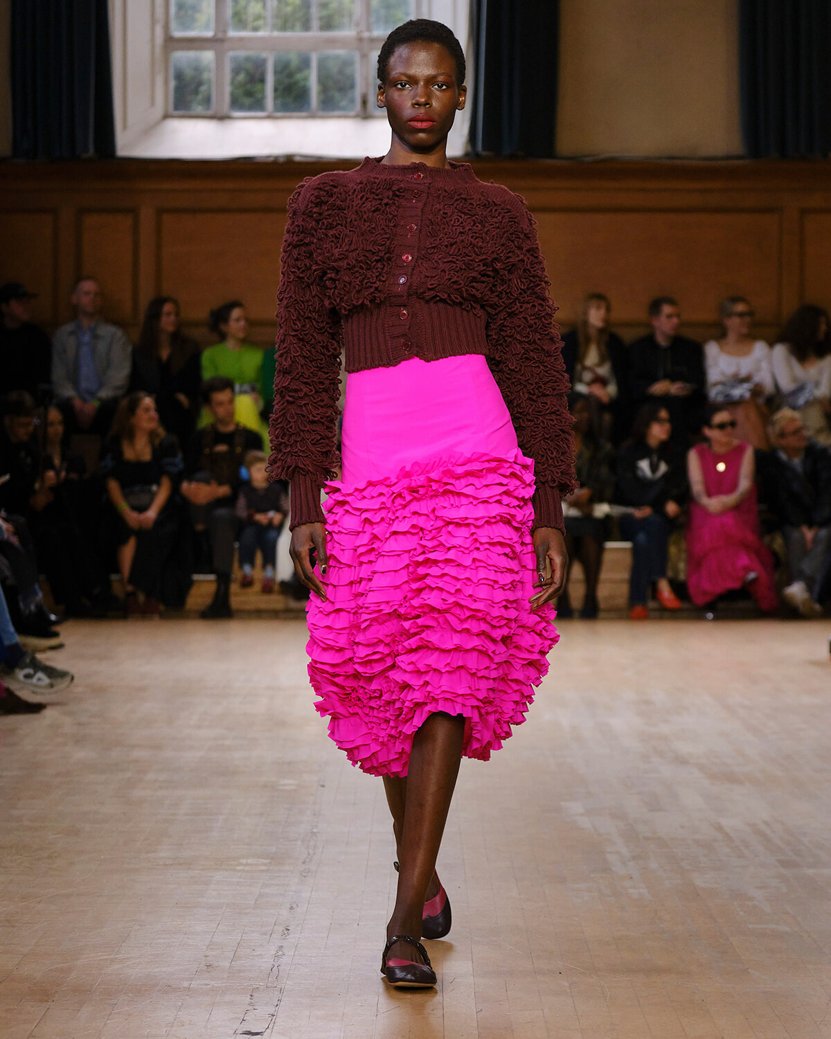 Model walks the runway in a burgundy textured cardigan with a bright pink, layered ruffled skirt; audience sits along the sides in a gallery-style room.