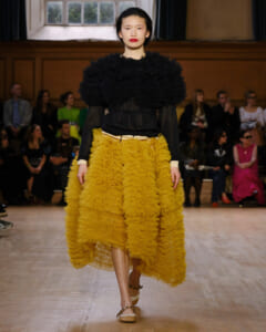 Model walks the runway in a black ruffled top and mustard-yellow layered skirt, audience watching in a dim venue.