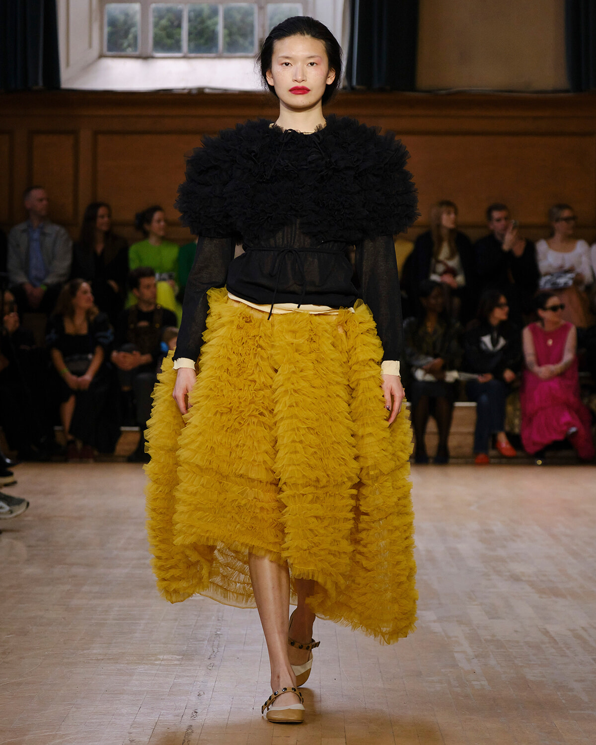 Model walks the runway in a black ruffled top and mustard-yellow layered skirt, audience watching in a dim venue.
