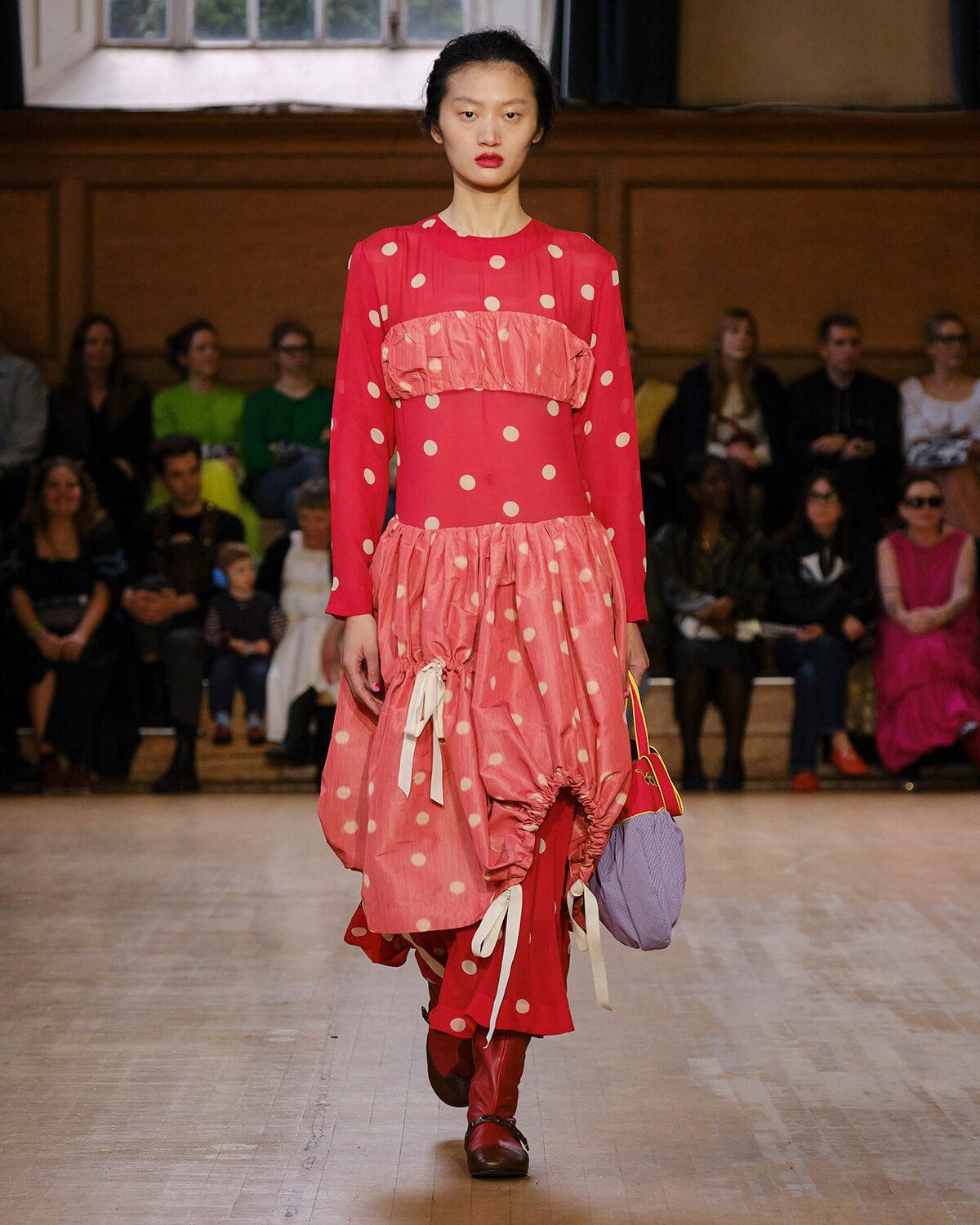 Model walks the runway in a bright pink polka-dot dress with ruffled panels and a lavender purse, audience in background.