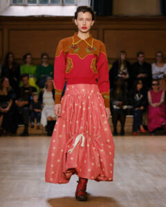Model on a runway wearing a burnt orange sweater with floral embroidery and a voluminous pink polka-dot skirt.