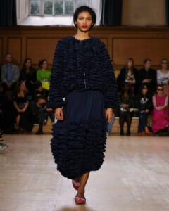 Model walks a runway in a navy textured ruffled outfit, audience seated along the sides in a warmly lit venue.