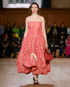 Model walking the runway in a pink strapless polka-dot dress with a voluminous ruffled skirt, holding a red handbag as spectators watch.