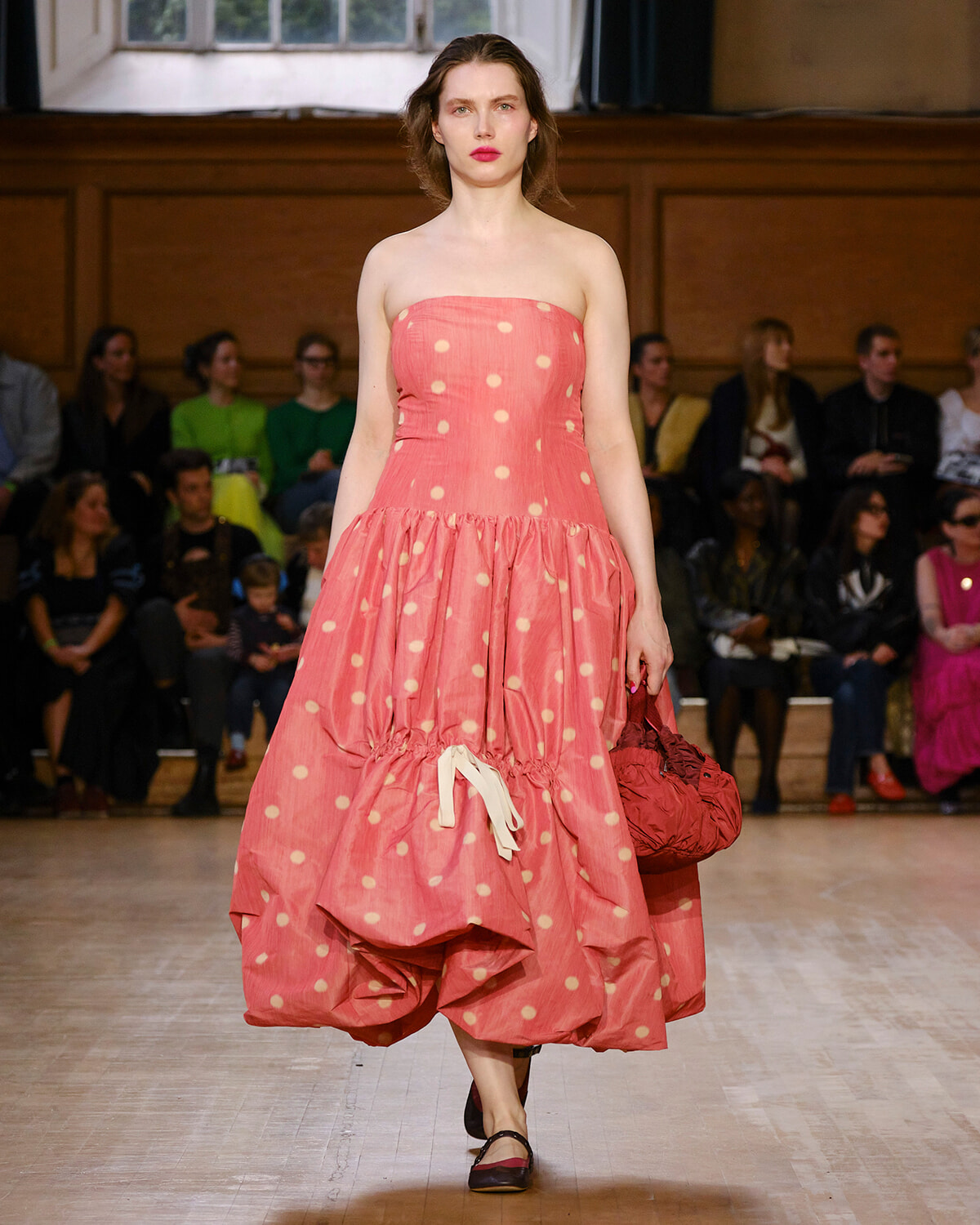 Model walking the runway in a pink strapless polka-dot dress with a voluminous ruffled skirt, holding a red handbag as spectators watch.