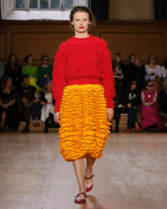 Model walks the runway in a red textured sweater and a bright orange ruffled midi skirt, with an audience in the background.