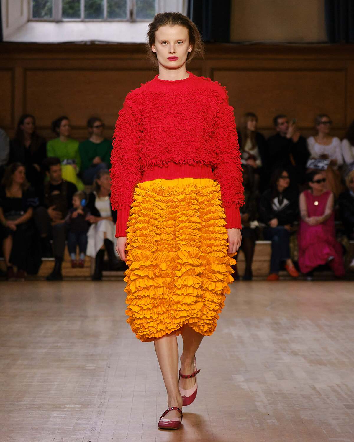 Model walks the runway in a red textured sweater and a bright orange ruffled midi skirt, with an audience in the background.