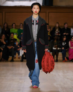 Model walking a runway in a long black coat with red piping, a gray-embellished shirt, blue jeans, and a red ruched handbag.