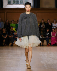 Model walking a fashion show in a chunky gray knit sweater with flower embellishments and a cream tulle skirt; audience in background.